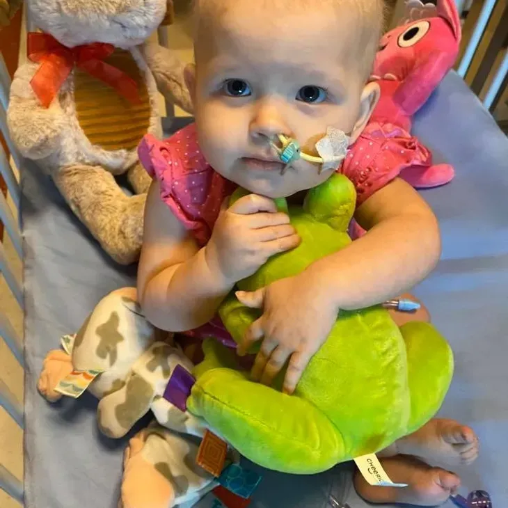 Jazmine holding a big green plush frog in her crib, surrounded by her favorite soft toys.