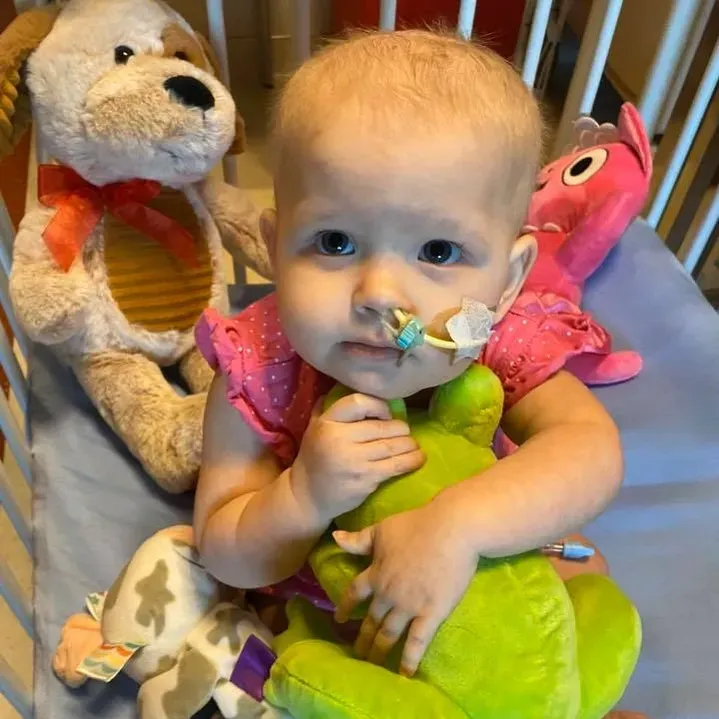 Jazmine in a pink dress, holding a big green plush frog close in her hospital crib.