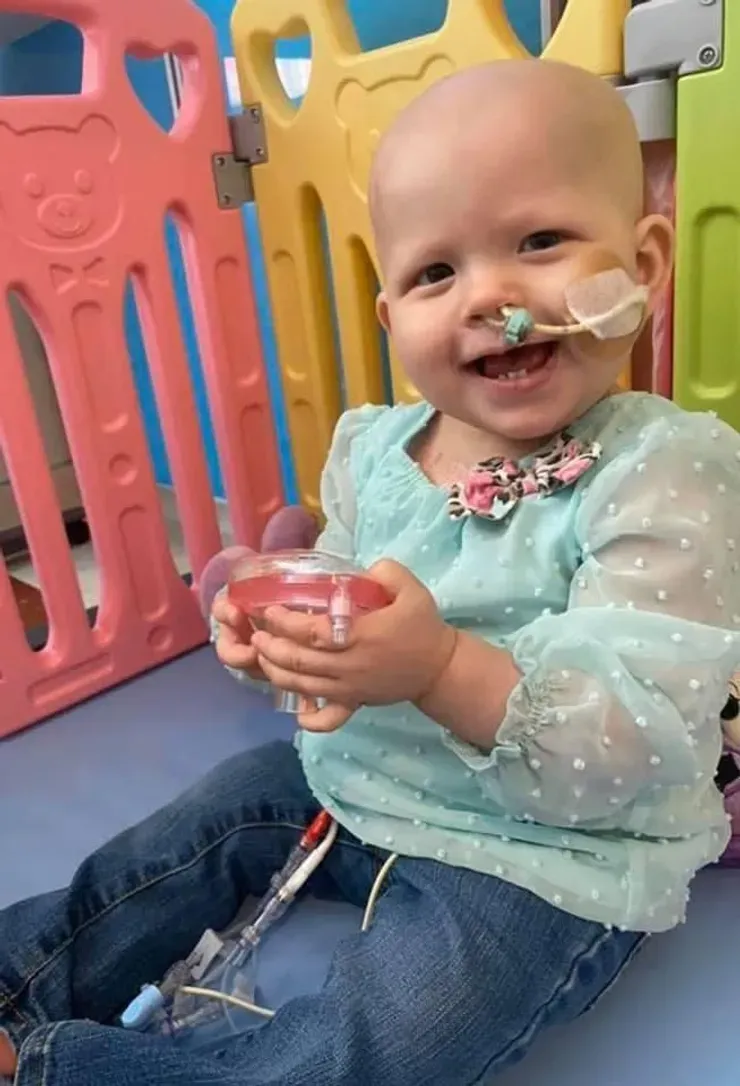Jazmine sitting up in a bright playroom, grinning as she holds a small medicine cup.