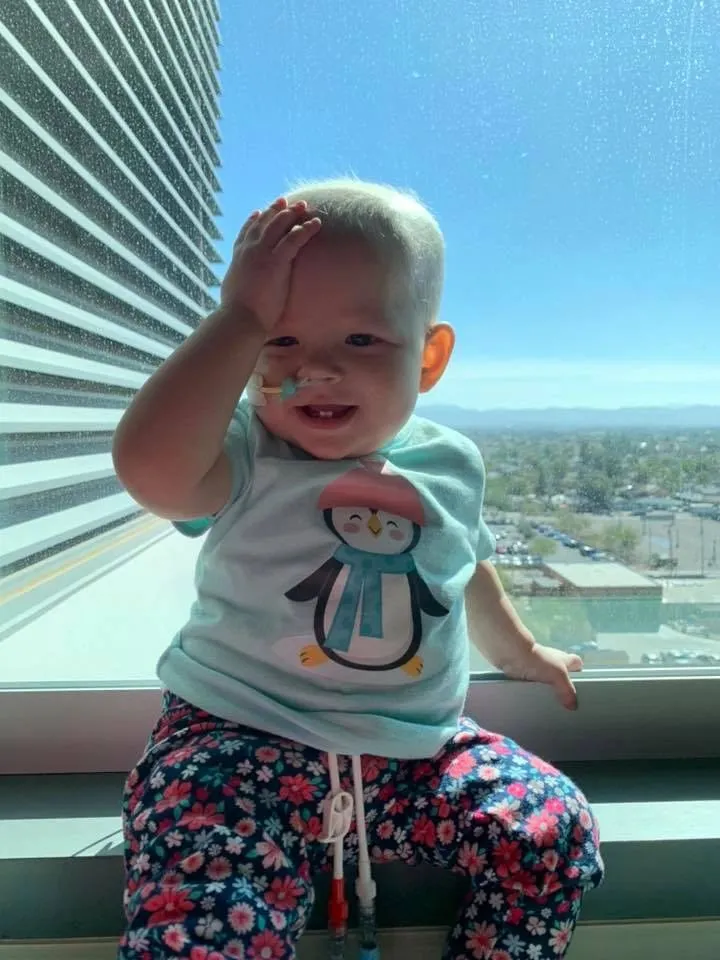 Jazmine sitting on a sunlit hospital windowsill, beaming, one hand raised to her forehead.